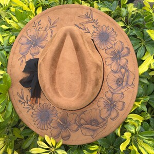 May include: A brown fedora hat with a floral design. The hat has a wide brim and a teardrop-shaped crown. The brim is decorated with a dark floral pattern. A black ribbon and tassel are tied around the base of the crown.
