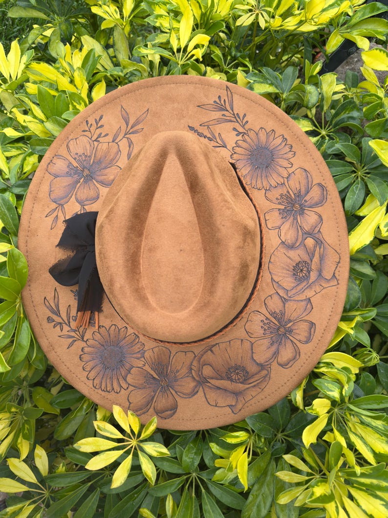 May include: A brown fedora hat with a floral design. The hat has a wide brim and a teardrop-shaped crown. The brim is decorated with a dark floral pattern. A black ribbon and tassel are tied around the base of the crown.