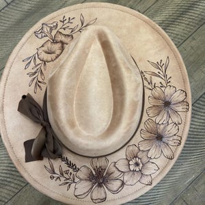 May include: A beige fedora hat with a floral design. The hat has a brown ribbon and a detailed pattern of flowers and leaves around the brim. The hat is a neutral colour, suitable for various outfits.
