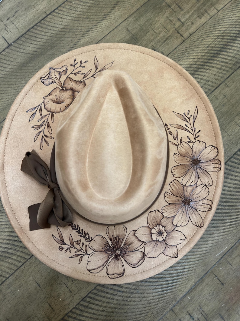 May include: A beige fedora hat with a floral design. The hat has a brown ribbon and a detailed pattern of flowers and leaves around the brim. The hat is a neutral colour, suitable for various outfits.