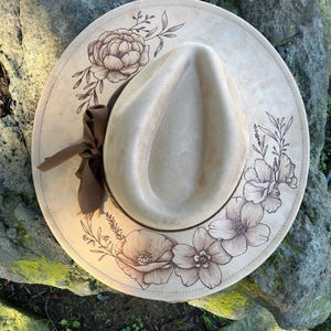 May include: A beige fedora hat with a wide brim, decorated with a brown floral design. The hat has a brown ribbon and is set against a natural background. The brim is decorated with detailed drawings of flowers and leaves.