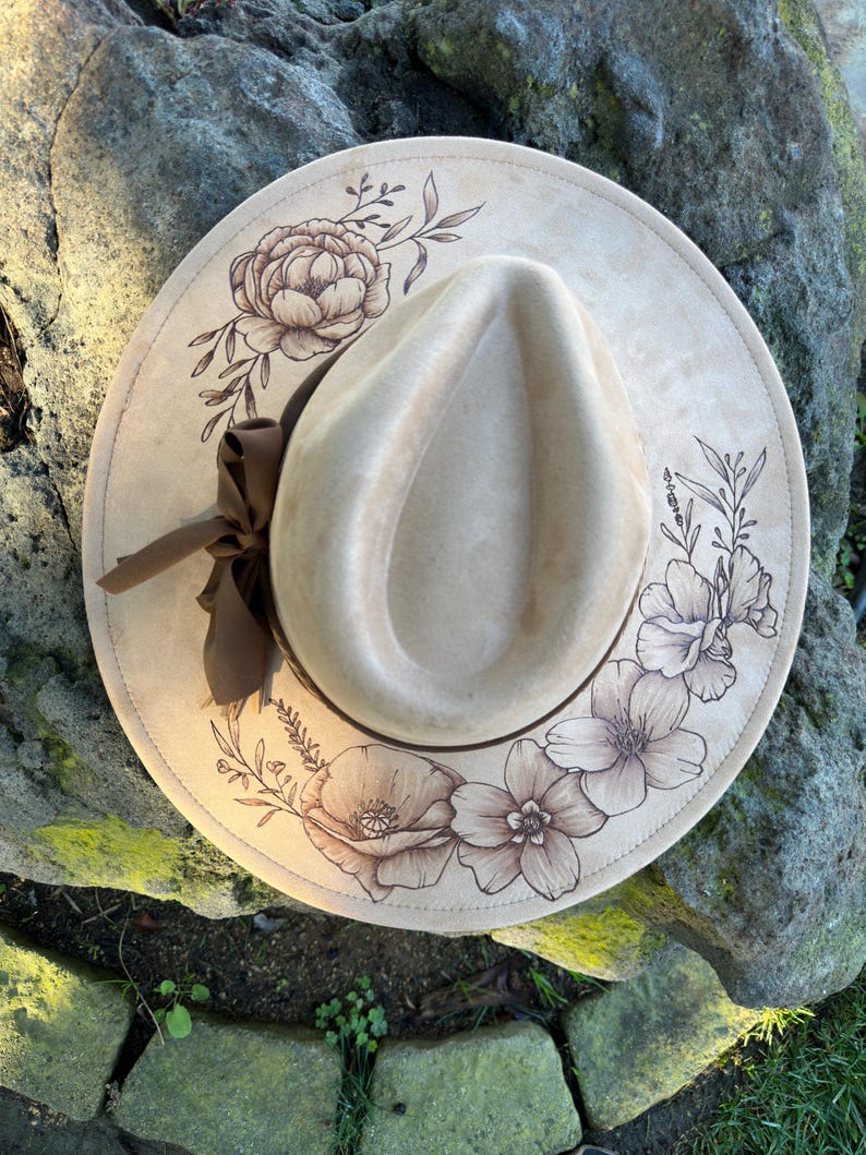 May include: A beige fedora hat with a wide brim, decorated with a brown floral design. The hat has a brown ribbon and is set against a natural background. The brim is decorated with detailed drawings of flowers and leaves.