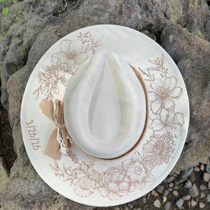 May include: Cream-coloured fedora hat featuring a light brown floral design. The brim is adorned with various flowers and leaves. A small bow and lace are attached to the side. The date 3/26/26 is printed on the brim.