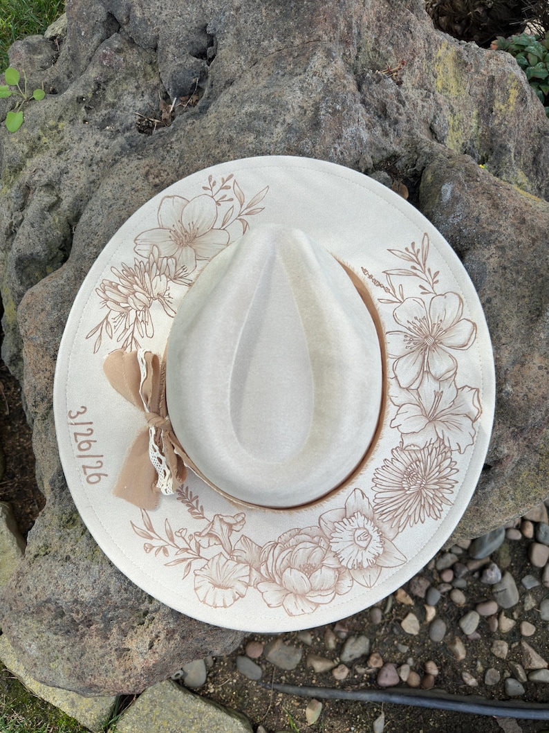 May include: Cream-coloured fedora hat featuring a light brown floral design. The brim is adorned with various flowers and leaves. A small bow and lace are attached to the side. The date 3/26/26 is printed on the brim.