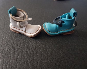 Puede incluir: Dos llaveros con forma de bota en miniatura, uno de cuero blanco y otro de cuero azul brillante. Cada bota cuenta con una anilla metálica sujeta a una trabilla en el talón, costuras visibles a lo largo de la suela y un pequeño detalle de cordones.