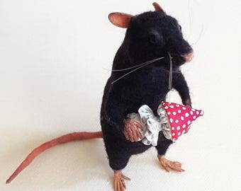 May include: A needle-felted black rat figurine standing upright. The rat has a long pink tail, pink ears, and long whiskers. It is holding a small, ruffled white fabric collar and a red party hat with white polka dots in its front paws.