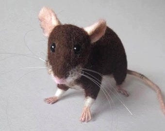 May include: A needle-felted mouse figurine with dark brown fur, white paws, and a pink nose. The mouse features black bead eyes, long white whiskers, and large ears with light pink inner coloring, standing on a light gray surface.