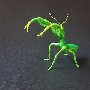 May include: A small, handcrafted sculpture of a praying mantis in a vibrant lime green colour. The figure features long, thin legs, a curved body, and two raised, spiky forelimbs. It has a textured surface with subtle yellow highlights on the head.