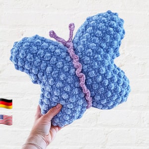 May include: A blue crocheted butterfly pillow with a pink crocheted center. The pillow is being held by a hand.