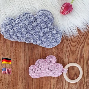 Può includere: Due giocattoli a forma di nuvola all'uncinetto. Uno è una nuvola viola chiaro con un anello di legno attaccato. L'altro è una nuvola viola scuro.