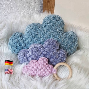 Può includere: Tre cuscini a forma di nuvola all'uncinetto in blu, viola e rosa, con un anello da dentizione in legno. I cuscini sono realizzati con un filato testurizzato e sono disposti in ordine decrescente di dimensioni.