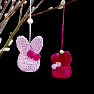Könnte beinhalten: Zwei gehäkelte Hasen-Anhänger, die an einem Zweig hängen. Der Hase links ist helllila mit einer rosa Schleife. Der Hase rechts ist rosa mit einer helllila Schleife.
