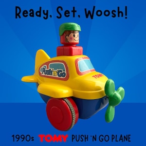 May include: A vintage yellow, blue, and red Tomy Push 'n Go plane toy from the 1990s. The toy plane has a green propeller, red wheels, and a small pilot figure. Text on the plane reads "Pushin' Go" and "Ready, Set, Woosh!"