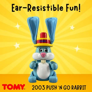 May include: A blue and white toy rabbit with large blue ears and a red and yellow hat. The toy has a white belly and a pink nose. The text "Ear-Resistible Fun!" is at the top, and "TOMY" and "2003 PUSH 'N GO RABBIT" are at the bottom.