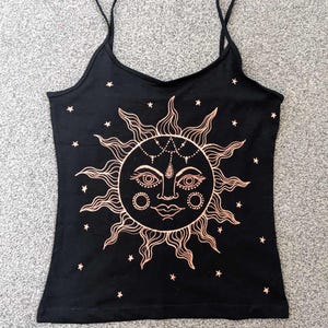 May include: A black tank top featuring a gold sun and stars design. The sun has a face with a teardrop and a small circle on each cheek.