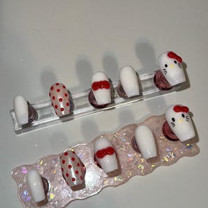 Puede incluir: Un conjunto de uñas postizas con varios diseños. Algunas uñas son blancas lisas, otras tienen lunares rojos y algunas están decoradas con un lazo rojo. Dos uñas están diseñadas con una cara de Hello Kitty, con un lazo rojo.