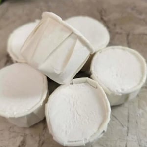 May include: A group of six white paper cups filled with a white substance, possibly frosting or a similar confection. The cups are arranged in a semi-circle on a light gray surface.