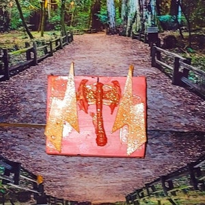 May include: A pink resin mold with a gold glitter finish. The mold is shaped like a double-headed axe with wings. The mold is sitting on a dirt path in a forest.