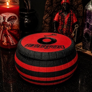 May include: A round, lidded container featuring alternating horizontal black and red textured stripes. The lid displays the text "ELEGUA" in a bold, stylized font, accompanied by a small black key icon centered above the lettering.