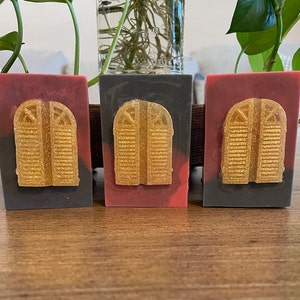 May include: Three rectangular soaps with a gold-colored, glittery door design. The soaps have a gradient of colors, with a dark gray base, a red middle, and a black top. The soaps are displayed on a wooden surface.