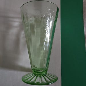 May include: A vintage green glass sundae or parfait glass. The glass has a flared cone shape with etched floral designs near the rim. The base is round with vertical ridges. The glass is approximately 7 inches tall.