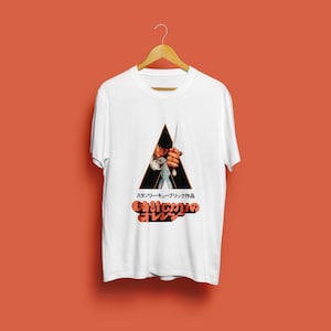 Pode incluir: T-shirt branca com um gráfico do filme "Laranja Mecânica". O design inclui uma imagem triangular de um personagem segurando uma lâmina, com texto japonês abaixo. A camisa está pendurada em um cabide de madeira contra um fundo laranja.