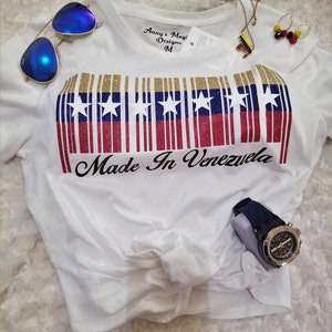 Puede incluir: Camiseta blanca con un diseño de código de barras de purpurina dorada que presenta los colores de la bandera venezolana y estrellas. El texto "Made in Venezuela" está impreso debajo del código de barras. La camiseta está anudada en la parte delantera.