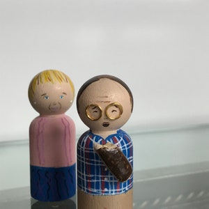 Puede incluir: Dos mu&ntilde;ecos de madera, uno con cabello rubio y una camisa rosa, el otro con cabello casta&ntilde;o, gafas y una camisa a cuadros azules sosteniendo un objeto marr&oacute;n.