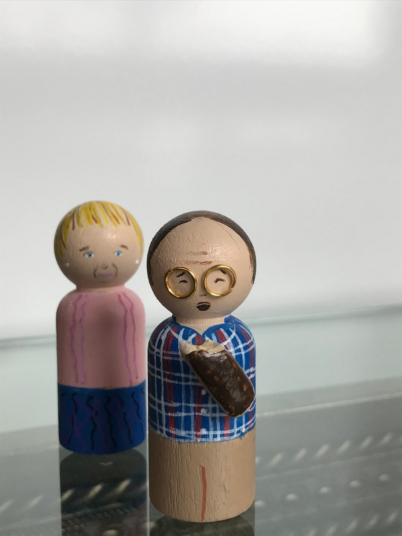 Puede incluir: Dos mu&ntilde;ecos de madera, uno con cabello rubio y una camisa rosa, el otro con cabello casta&ntilde;o, gafas y una camisa a cuadros azules sosteniendo un objeto marr&oacute;n.