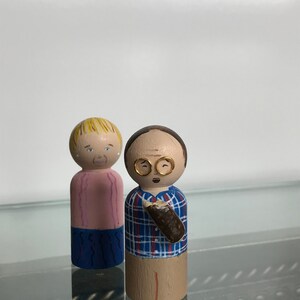 Puede incluir: Un par de mu&ntilde;ecas de madera pintadas a mano. La mu&ntilde;eca femenina tiene el pelo rubio, una camisa rosa y pantalones azules. La mu&ntilde;eca masculina tiene el pelo casta&ntilde;o, gafas, una camisa a cuadros azul y roja y pantalones marrones. &Eacute;l sostiene un objeto marr&oacute;n en su mano derecha.