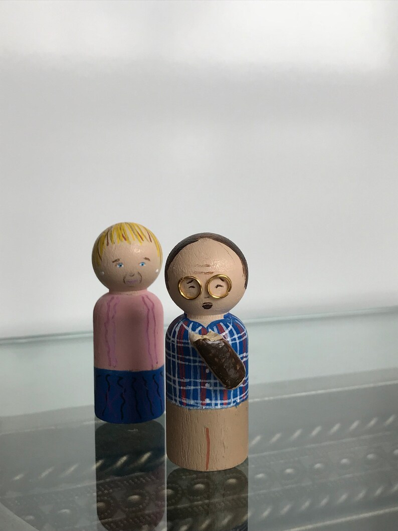 Puede incluir: Un par de mu&ntilde;ecas de madera pintadas a mano. La mu&ntilde;eca femenina tiene el pelo rubio, una camisa rosa y pantalones azules. La mu&ntilde;eca masculina tiene el pelo casta&ntilde;o, gafas, una camisa a cuadros azul y roja y pantalones marrones. &Eacute;l sostiene un objeto marr&oacute;n en su mano derecha.