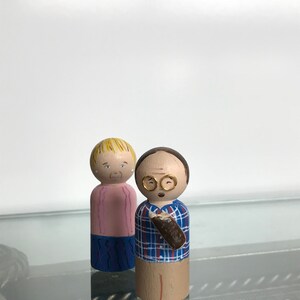 Puede incluir: Dos mu&ntilde;ecas de madera con clavijas, una con cabello rubio que lleva una camisa rosa y pantalones azules, la otra con cabello casta&ntilde;o que lleva una camisa a cuadros azules y blancos y sostiene un objeto marr&oacute;n.