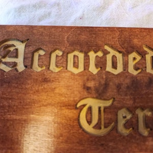 以下が含まれることがあります： 「Accorded」と「Terr」という文字が金色の文字で彫られた木製の看板です。