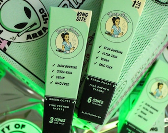 May include: Several boxes of green cones are displayed, featuring the "Blazy Susan" logo with a woman in a blue dress. The boxes are labeled "King Size" and "1 1/4 Size" and list features like "Slow Burning" and "Vegan."