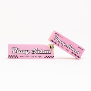 May include: Two pink boxes of Blazy Susan 1/4 size rolling papers. The boxes are pink with black lettering and a checkerboard pattern. The text on the boxes reads "Blazy Susan" and "Pink Rolling Papers".