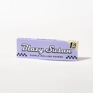 May include: A lavender box of "Blazy Susan" purple rolling papers. The box has black and white checkered accents and the text "50 LEAVES PER PACK" and "1 1/4 SIZE". The brand name is in a bold, stylized font.