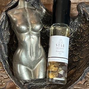 May include: A clear glass bottle with a black rollerball top filled with a golden liquid. The bottle is labeled "STAR AMETHYSTINE Ritual Oil 10ml Moon Time". The bottle is sitting on a silver metal figurine of a woman's torso.