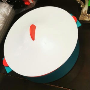 Puede incluir: Vaporera de silicona blanca con un borde azul turquesa y un asa roja. La vaporera tiene forma redonda y está diseñada para cocinar alimentos.