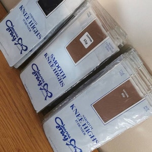 Puede incluir: Un paquete de medias Joanna Gray en negro, marrón y natural. Los paquetes están etiquetados con "Smooth Knee Highs 15 Denier" y muestran el logotipo de Joanna Gray. Los paquetes tienen un precio de 75p, 55p y 80p.