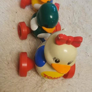 Może przedstawiać: Trzy kolorowe zabawki w kształcie kaczki do ciągnięcia. Kaczki są żółte, zielone i białe z czerwonymi kołami i akcentami. Biała kaczka ma czerwoną kokardę i napis "TOMY" na brzuchu.