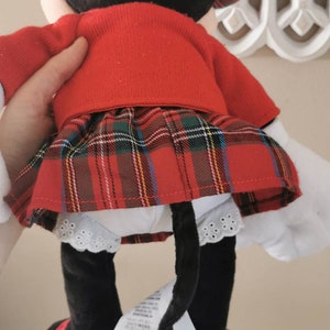 May include: A red and black plush toy of Minnie Mouse wearing a red sweater and a red and black plaid skirt. The toy has a white lace trim on the skirt and black shoes with red accents.