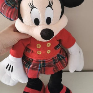 May include: A red and black plaid Minnie Mouse plush toy wearing a red dress with gold buttons and black shoes.