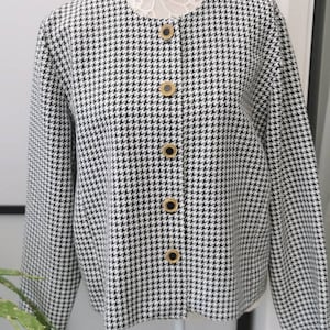 Puede incluir: Una chaqueta de punto de pata de gallo en blanco y negro con botones dorados. La chaqueta está en un maniquí blanco.