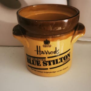 Puede incluir: Una pequeña olla de cerámica amarilla y marrón con asas, con el logotipo de Harrods y el texto "BLUE STILTON CHEESE". La olla tiene un borde marrón y probablemente es para servir queso.