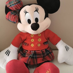May include: Red and black plaid Minnie Mouse plush toy wearing a red sweater with gold buttons and a red plaid skirt. The toy has a black bow with red and black plaid on its head. The toy's feet have the text "Disney Store 2016" embroidered on them.