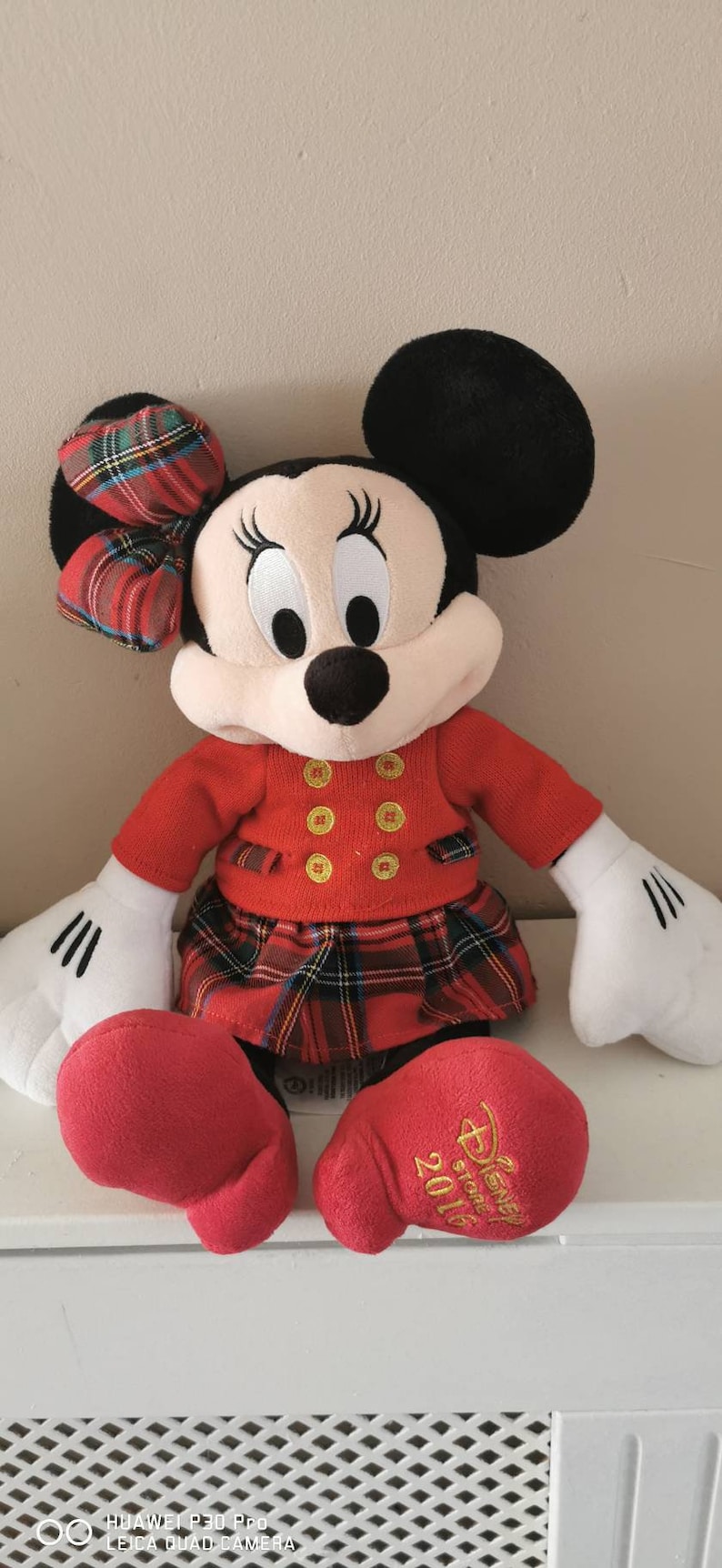 May include: Red and black plaid Minnie Mouse plush toy wearing a red sweater with gold buttons and a red plaid skirt. The toy has a black bow with red and black plaid on its head. The toy's feet have the text "Disney Store 2016" embroidered on them.