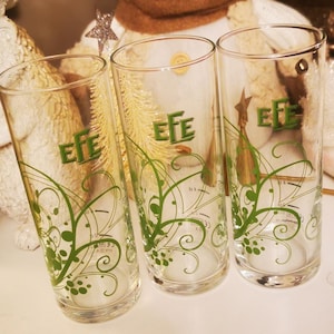 May include: Three clear glass tumblers, each with the word "EFE" in green and green floral patterns. A snowman decoration with a brown scarf and white hat is in the background. The glasses are on a white surface.
