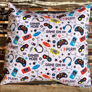 May include: A gray pillow with a repeating pattern of video game controllers, headphones, and the words "Play!" and "Game On!" The pattern is in black, red, blue, yellow, and white.