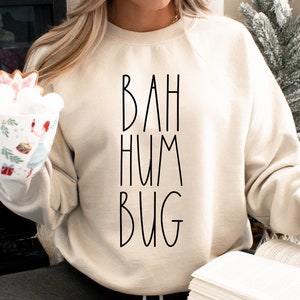 Puede incluir: Una sudadera de color crema con el texto "BAH HUM BUG" impreso en negro. La sudadera la lleva puesta una persona que sostiene una taza y un libro.