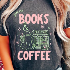May include: A charcoal gray short-sleeve t-shirt featuring a graphic print with the text "give me BOOKS & COFFEE" and "BOO-KSTORE". The design includes a ghost holding a coffee cup, a bookstore storefront, small flowers, and stars in pink and light green.